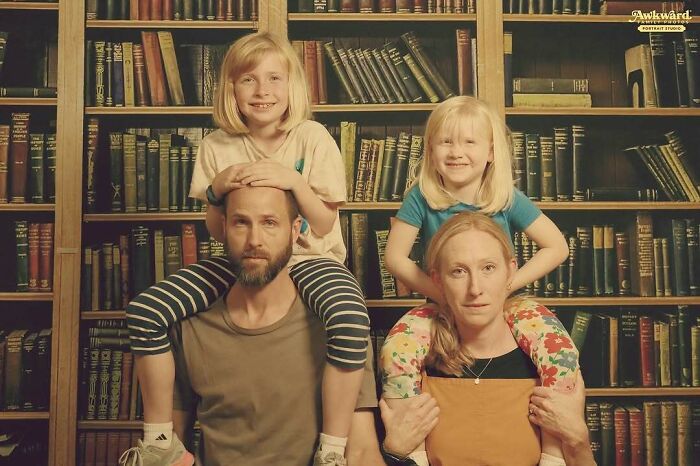 Family awkward studio photo with two children on parents' shoulders in front of a vintage bookshelf backdrop.