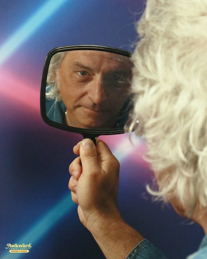 Man with curly white hair holding a mirror reflecting his face in an awkward studio photo setting with colorful backdrop.