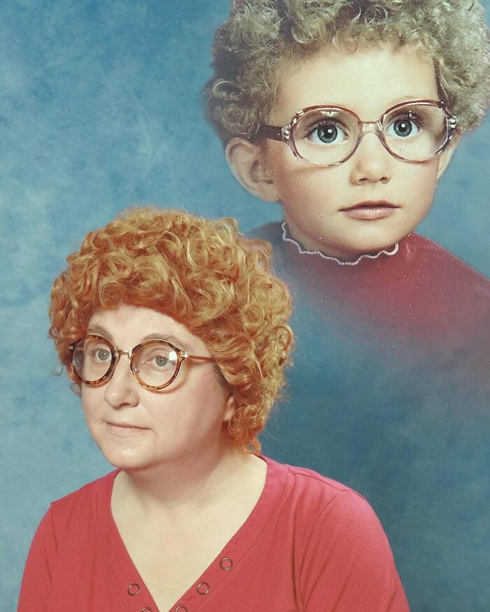 Woman with curly red wig and glasses posing for an awkward studio pic with a faded child portrait background.