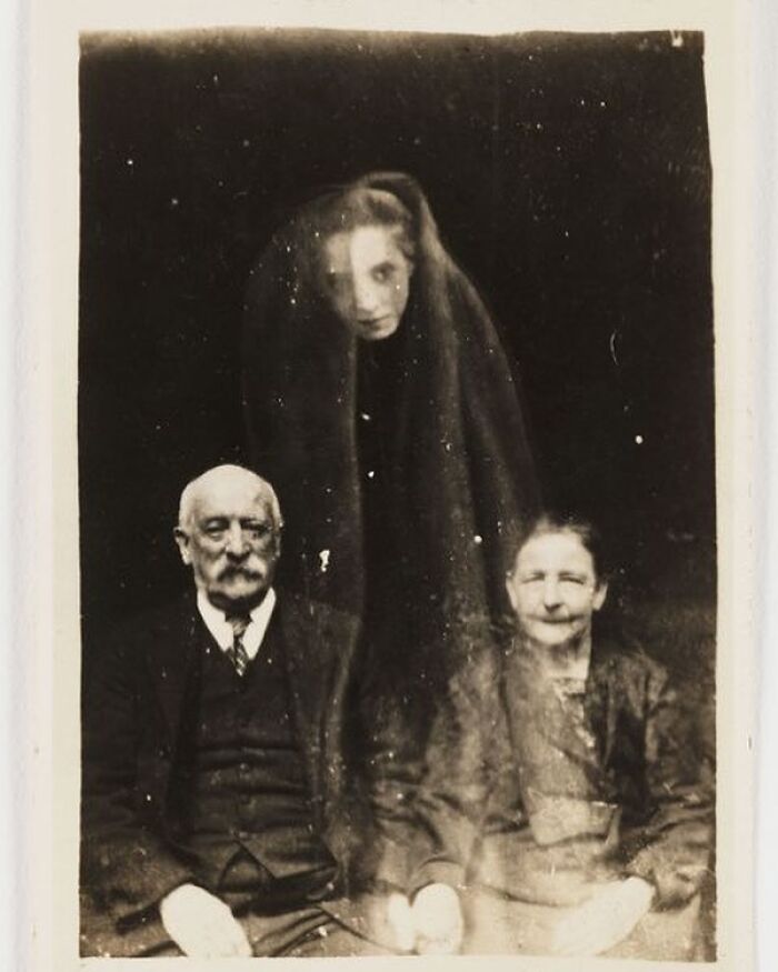Victorian era spirit photography showing an elderly couple with a ghostly figure behind them in a weird Victorian photo.