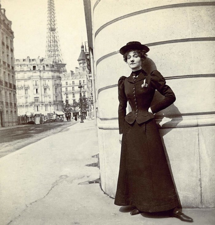 Victorian era woman in period dress posing against a curved building on a city street in a weird photo.