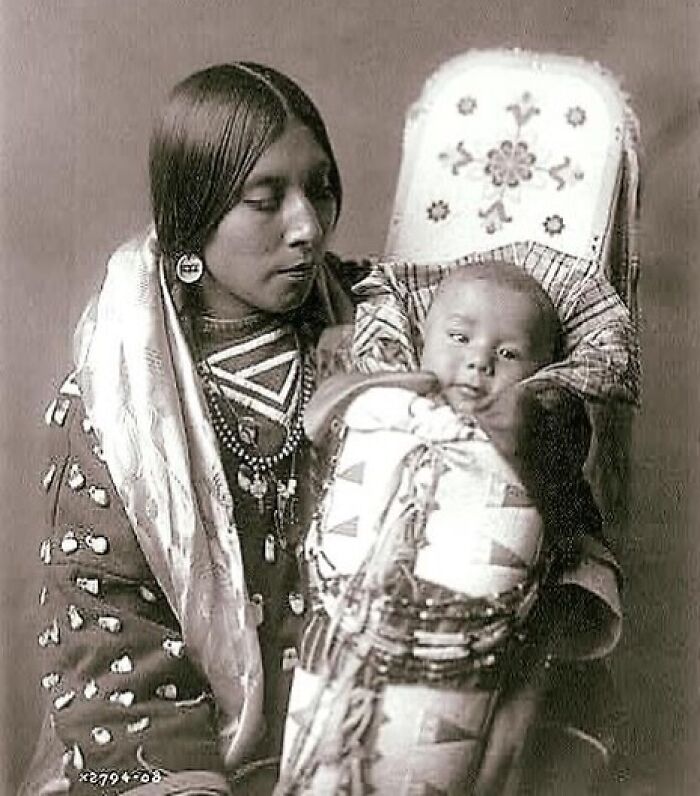 Victorian era photo of a Native American woman holding a wrapped baby in traditional clothing and beadwork.