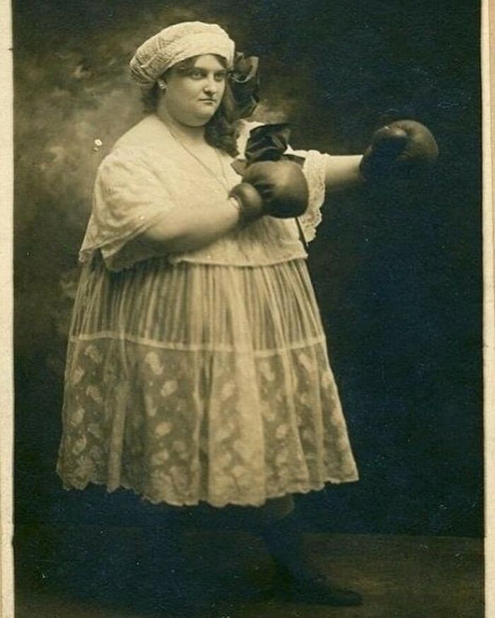Victorian era weird photo of a woman wearing boxing gloves and a lace dress posing in a vintage studio setting.
