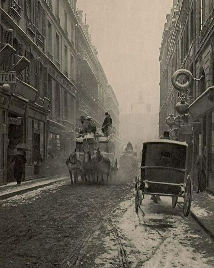 Victorian era street scene with horse-drawn carriages and pedestrians on a snowy, narrow city road.