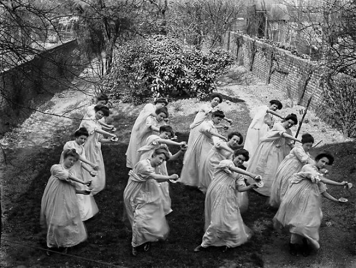 Group of Victorian-era women in long dresses performing a strange synchronized exercise in a garden setting.