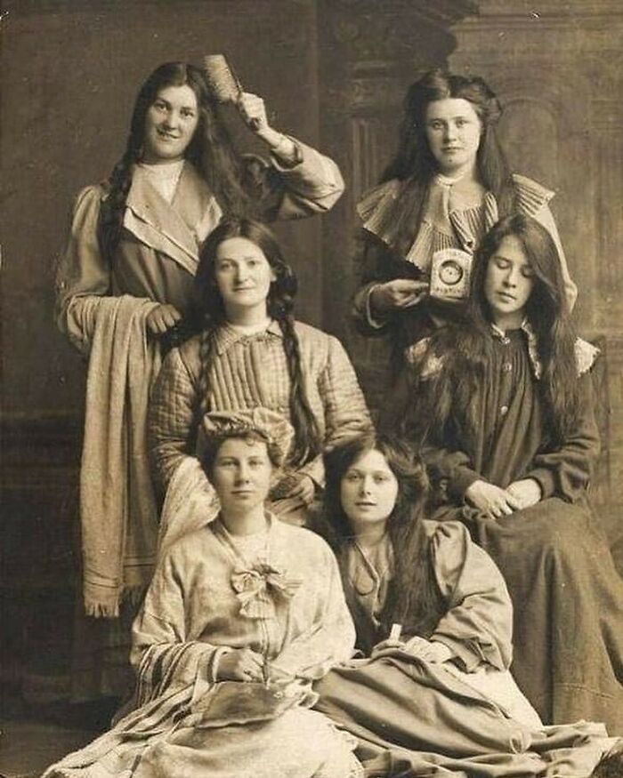 Six Victorian-era women with long hair pose in old-fashioned dresses in a weird photo from the Victorian era.