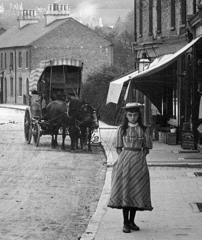 Victorian era street scene with a girl in period dress standing near a horse-drawn carriage in a small town setting.