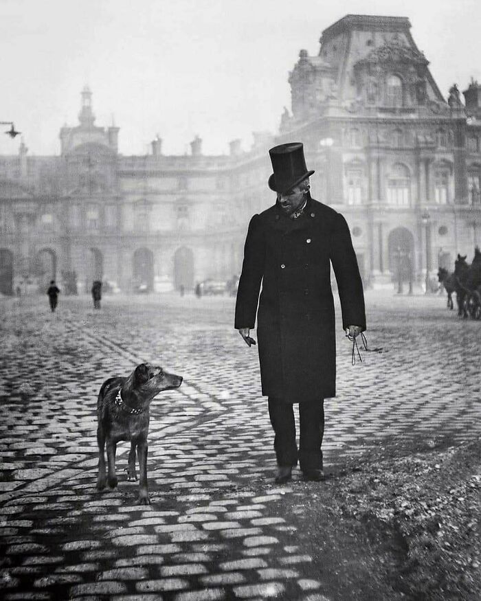 Victorian era man walking his dog on cobblestone street in front of historic building, capturing a weird photo moment.
