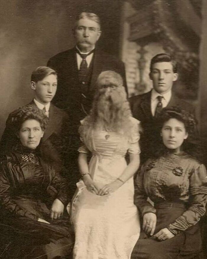 Victorian era weird photo of six people, including a woman with a full beard wearing a white dress, posing for a formal portrait.