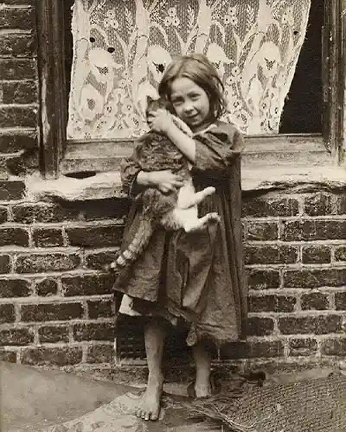 Victorian era black and white photo of a barefoot girl in a dress holding a cat outside a brick building with lace curtains.