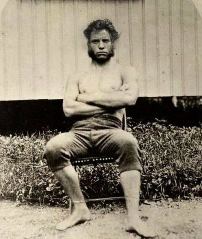 Victorian era weird photo of a shirtless man with large sideburns sitting barefoot on a chair outside.