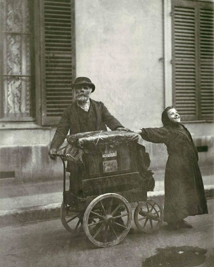 Victorian era photo of a man with a cart and a smiling woman holding hands on a city street in vintage clothing.