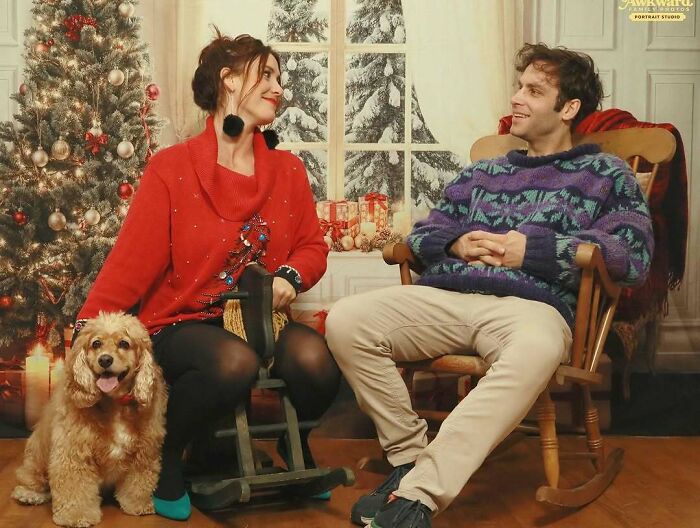 Couple in awkward studio photo with festive sweaters, vintage rocking chairs, and a dog in a holiday-themed setting.