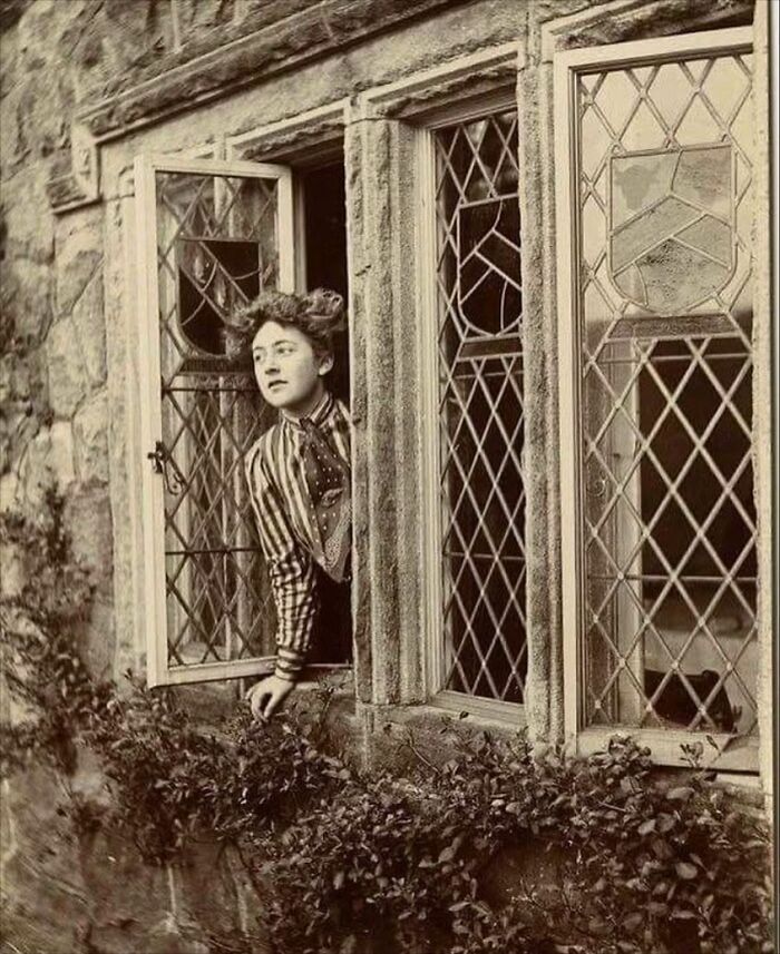 Young woman leaning out of a leaded glass window in a stone building in a weird Victorian era photo.