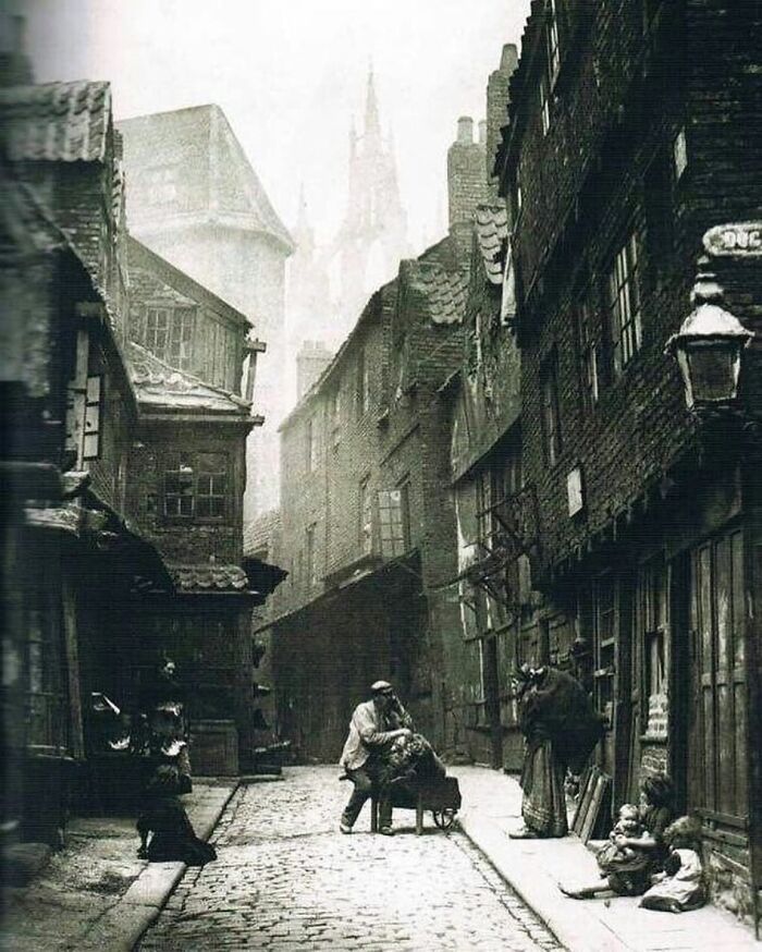 Victorian era street scene with cobblestone road, old buildings, and people engaged in daily life activities.