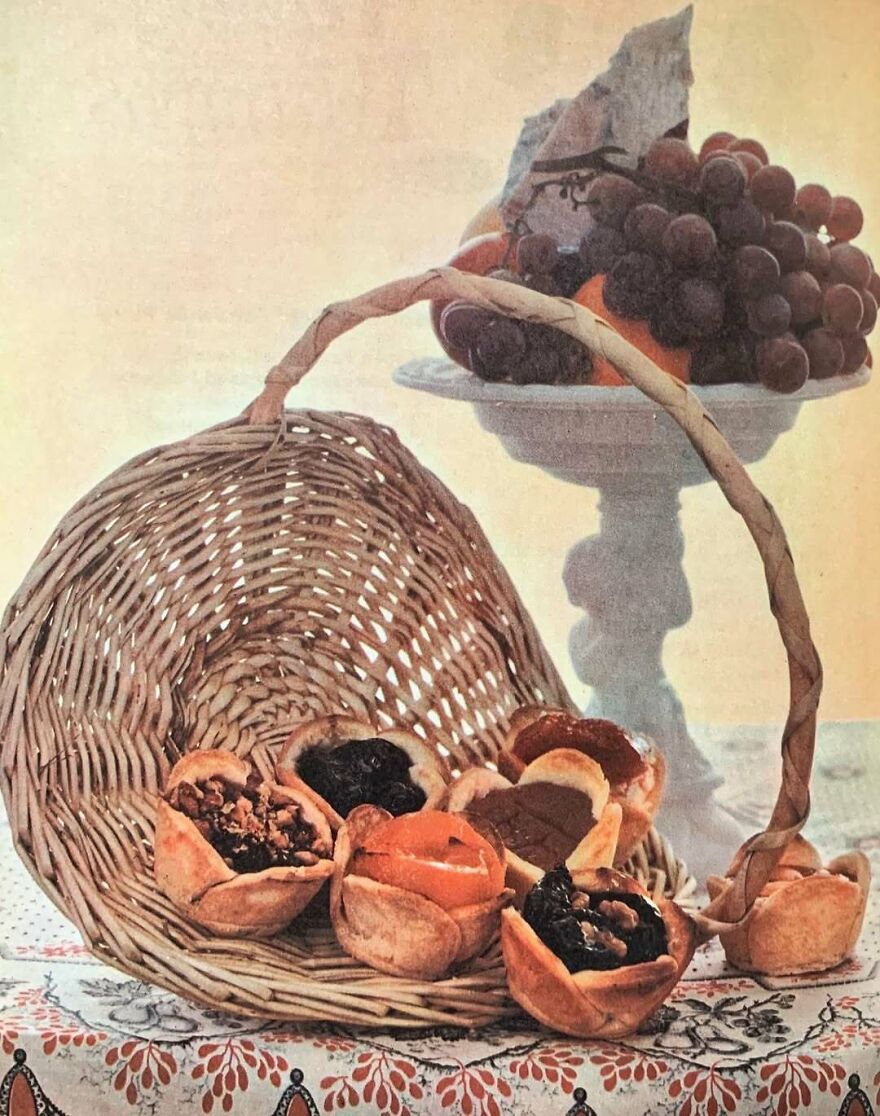 Wicker basket tipped over with vintage recipes dessert tarts on a patterned tablecloth and a fruit bowl in the background.