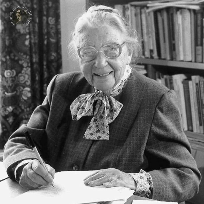 Elderly woman with glasses writing at a desk surrounded by books, representing overlooked women in world history achievements.