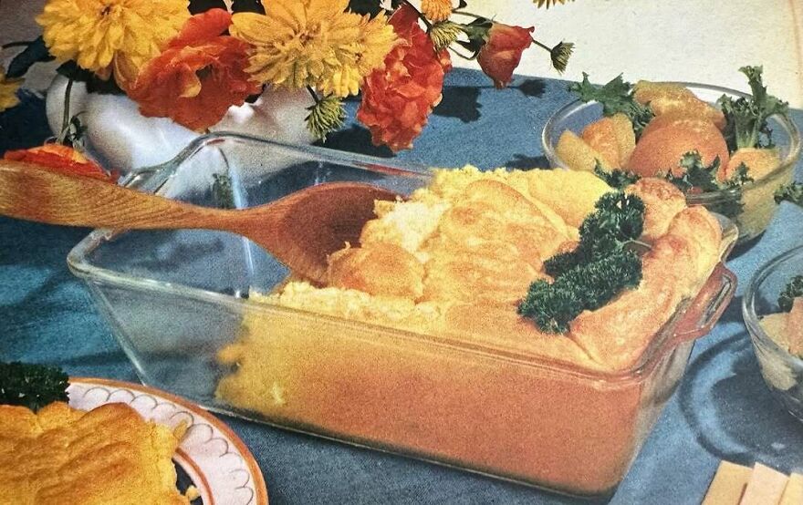 Vintage casserole dish with golden baked topping and parsley garnish surrounded by retro table setting and flowers