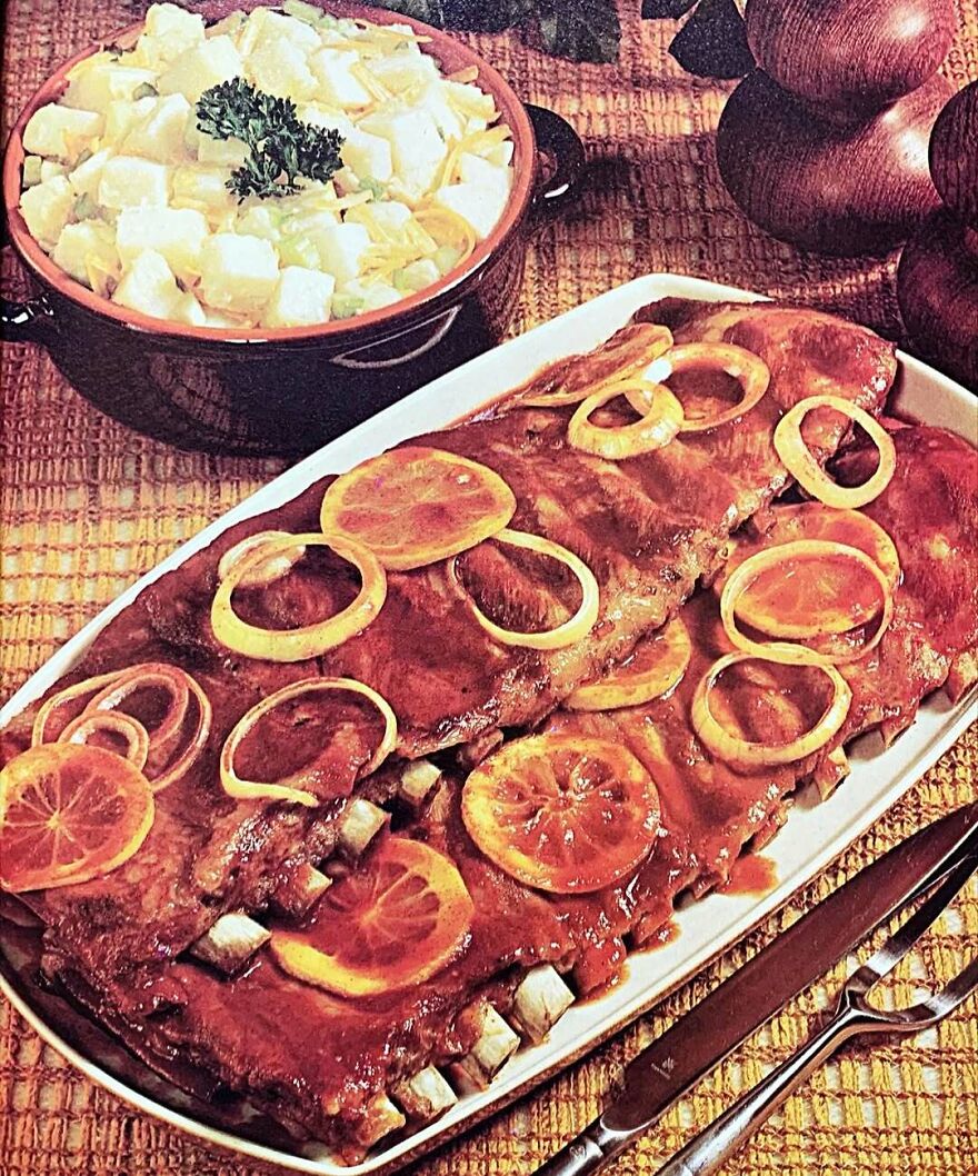 Vintage ribs covered with lemon slices and onions on a white platter alongside a bowl of creamy salad, showcasing vintage recipes.