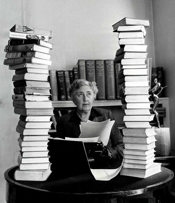 Black and white photo of a woman surrounded by towering stacks of books, symbolizing overlooked women in history achievements.