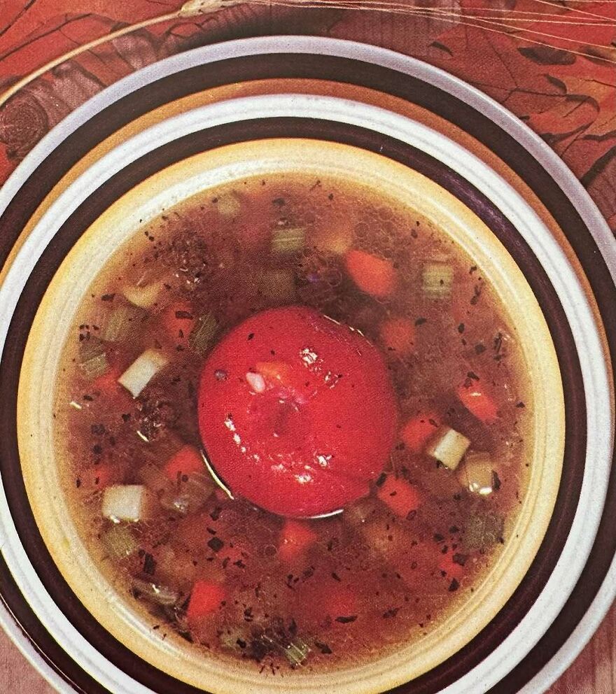 Bizarre vintage recipe of tomato-filled broth with diced vegetables in a white bowl on a patterned tablecloth.
