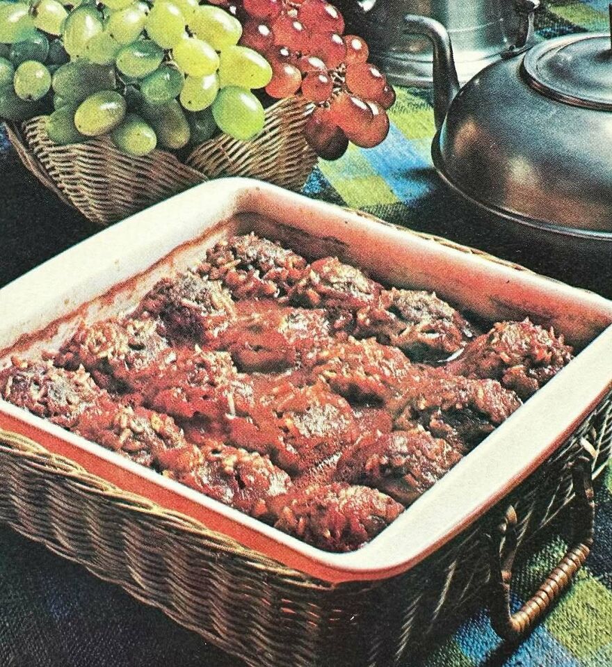 Vintage recipe casserole dish with a tomato-based sauce, grapes in a basket, and a metal kettle on a checkered tablecloth.