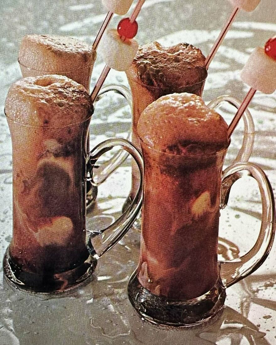 Four vintage recipes milkshake floats in glass mugs with skewered marshmallows and cherries on a wet surface.