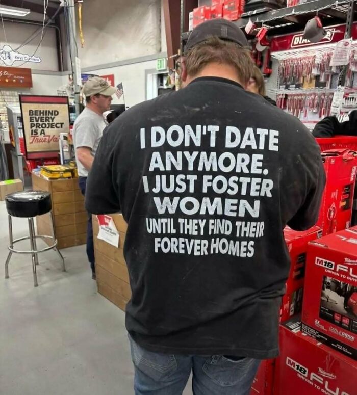 Man wearing a black shirt with a humorous quote, standing in a hardware store surrounded by tools and products, funny memes concept.