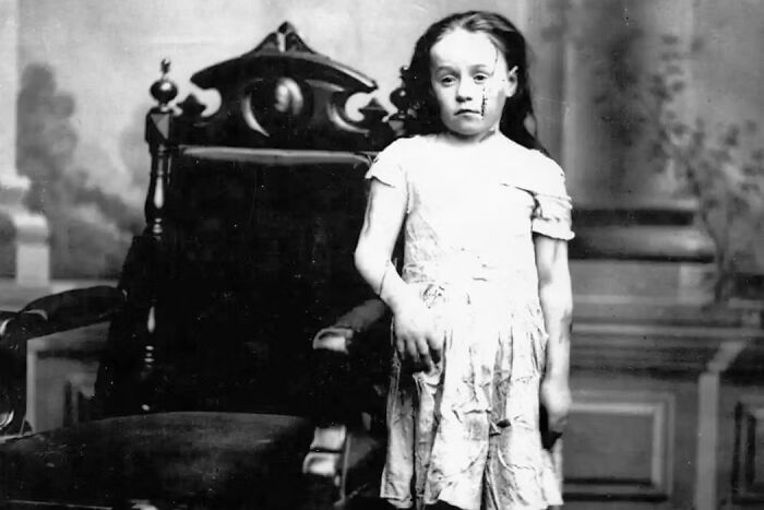 Black and white photo of a young girl standing beside an ornate chair, representing overlooked women in history and achievements.
