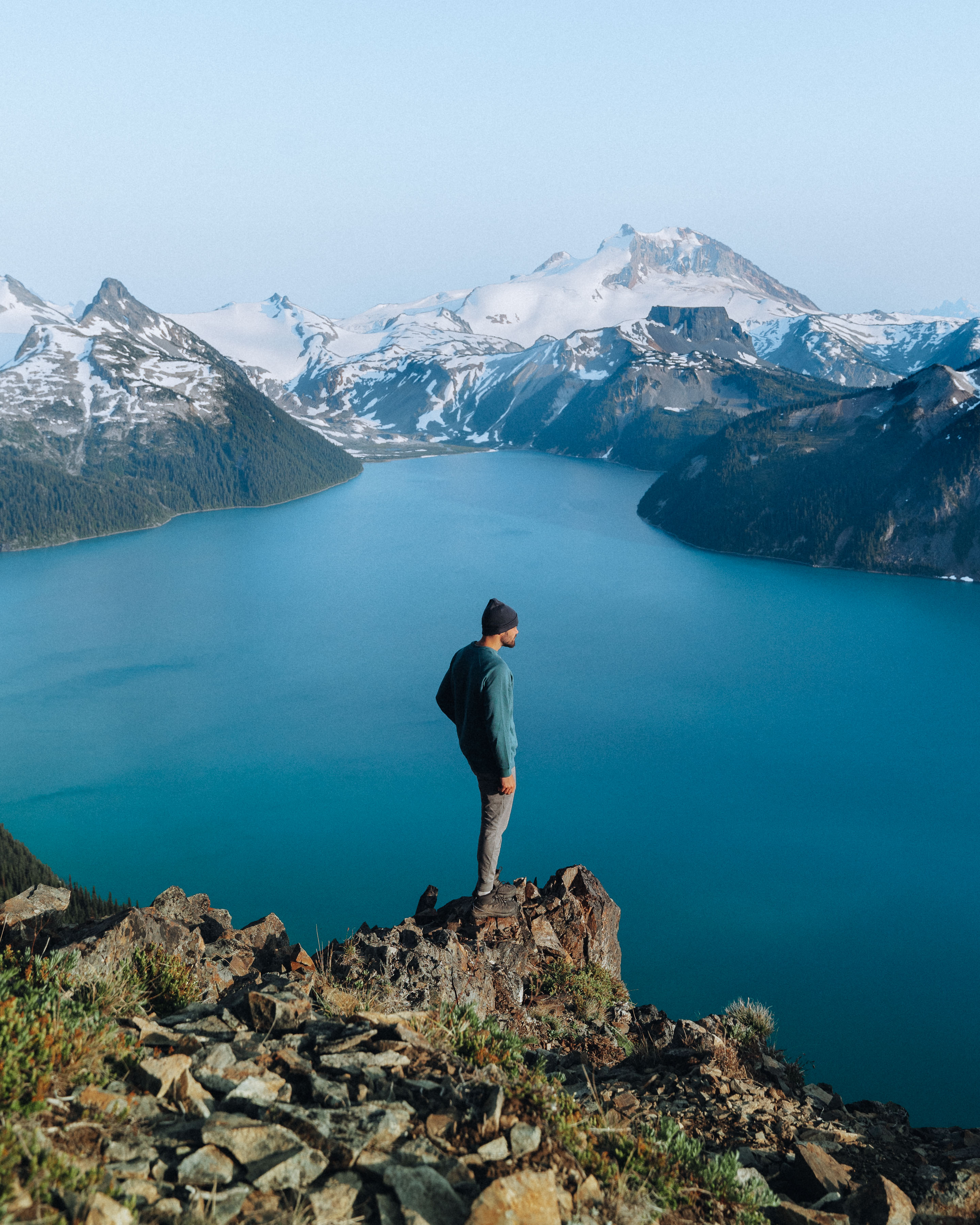 Man standing on rocky cliff overlooking a vast lake and snow-covered mountains, reflecting on purpose and journey. Man standing on rocky cliff overlooking a vast lake and snow-covered mountains, reflecting on purpose and journey.