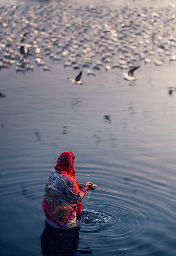 Woman in traditional clothing standing in water holding water with birds flying above in street photography around the world.