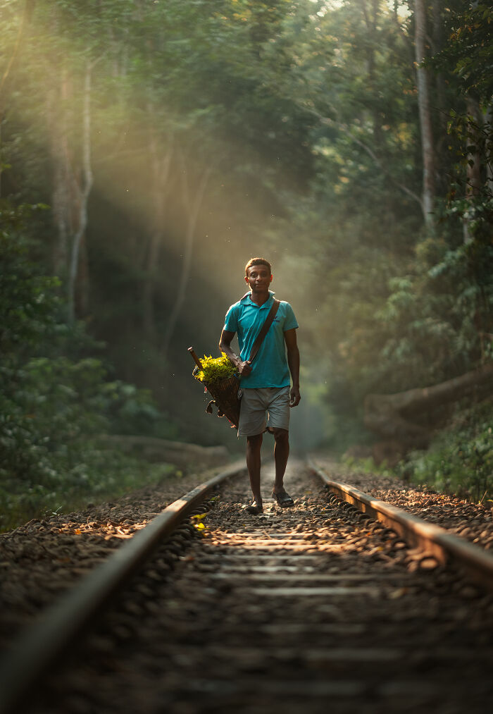 Young man carrying plants walking barefoot on railroad tracks in a forest, captured in street photography around the world.