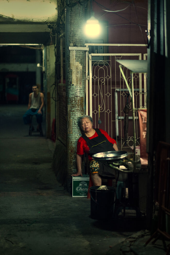 Elderly woman resting in a dimly lit alley, captured in street photography around the world with a moody urban atmosphere.