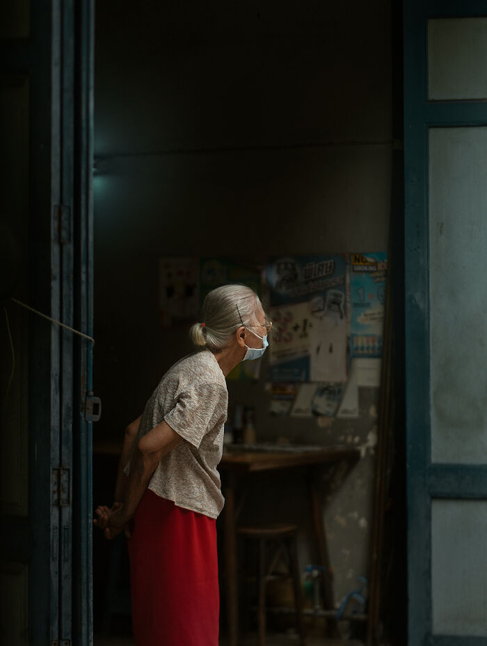 Elderly woman wearing a mask leaning forward inside a dimly lit room, captured in street photography around the world.