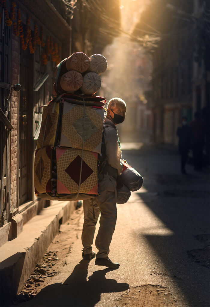 Man carrying large bundles on his back in a narrow street, captured in street photography with warm sunlight and shadows.