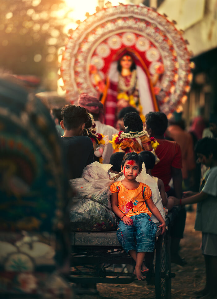 Young girl with traditional face paint sitting on a cart during a vibrant street photography scene in a cultural procession.