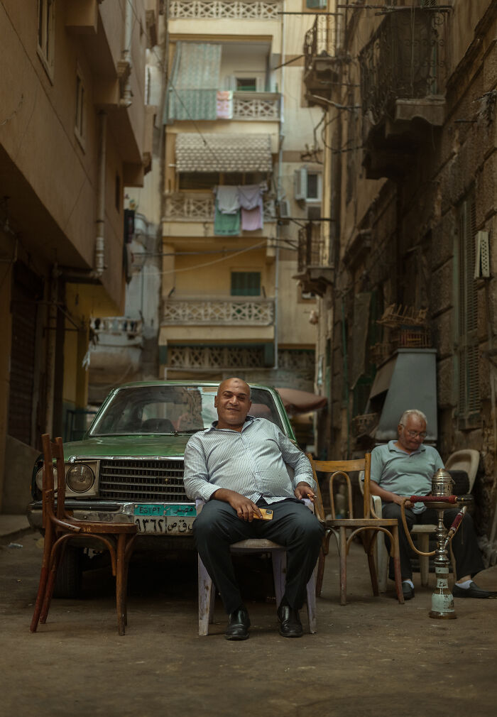 Two men sitting on chairs in a narrow alley with a green car, captured in street photography around the world.
