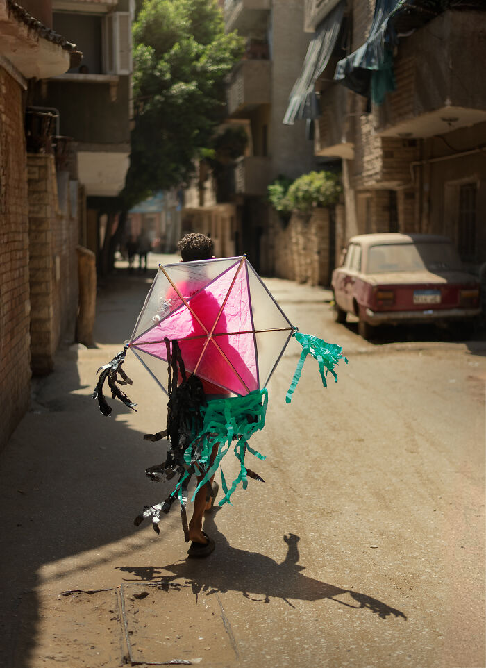 Child walking in a sunlit street holding a colorful kite, captured in a candid street photography shot.
