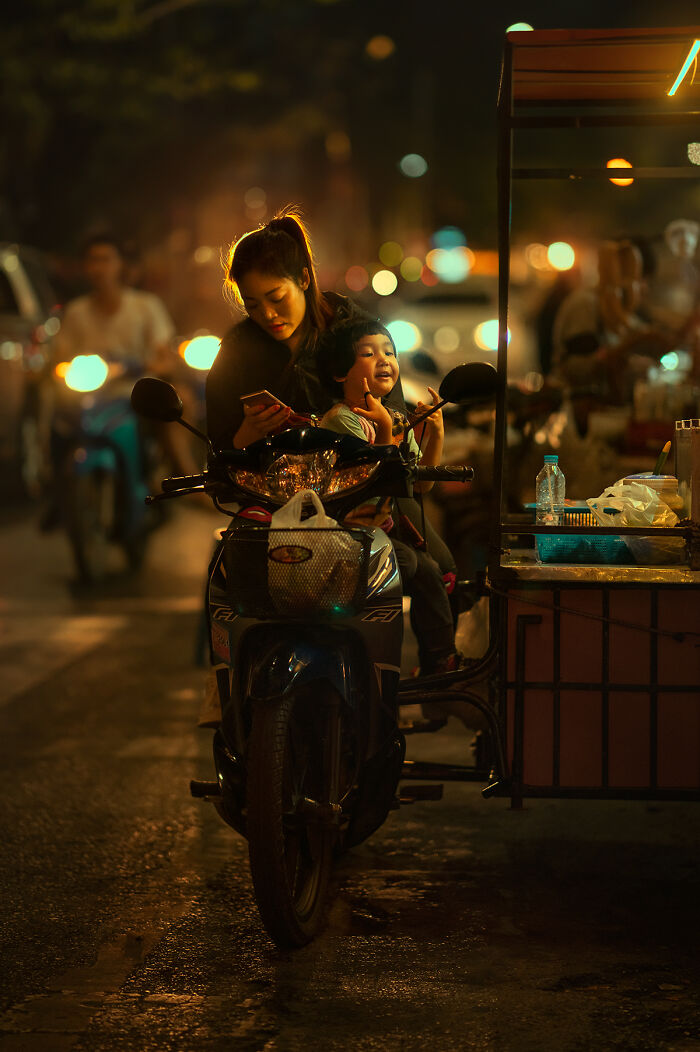 Young woman and child on a motorbike at night captured in street photography with vibrant urban lighting and bokeh effects.