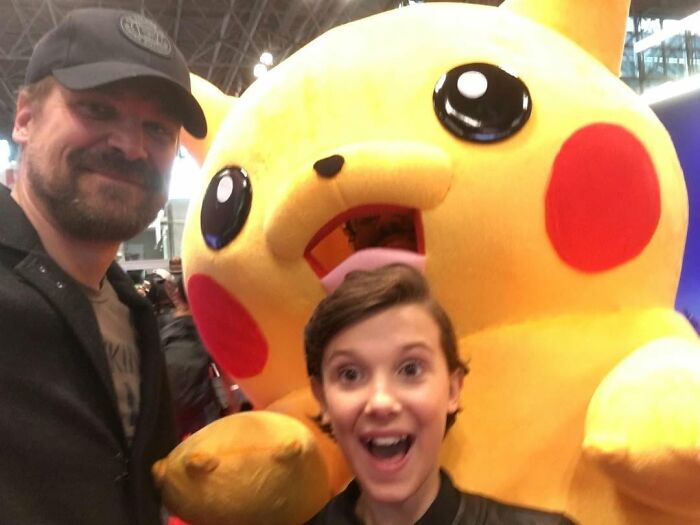Millie Bobby Brown and David Harbour posing happily with a large Pikachu mascot at a crowded event.