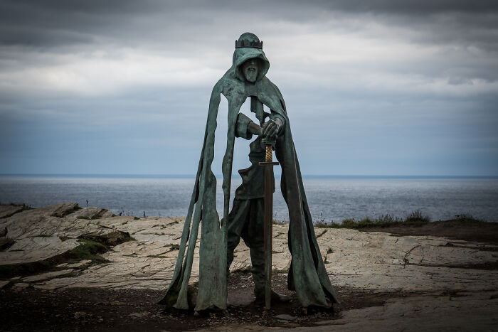 Bronze sculpture of a cloaked figure holding a sword, set against a rocky seaside backdrop, spectacular sculptures travel.