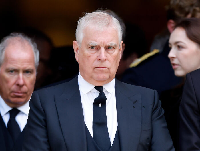Prince Andrew in a black suit and tie appearing somber, with focus on female touch influencing Andrew’s title removal.