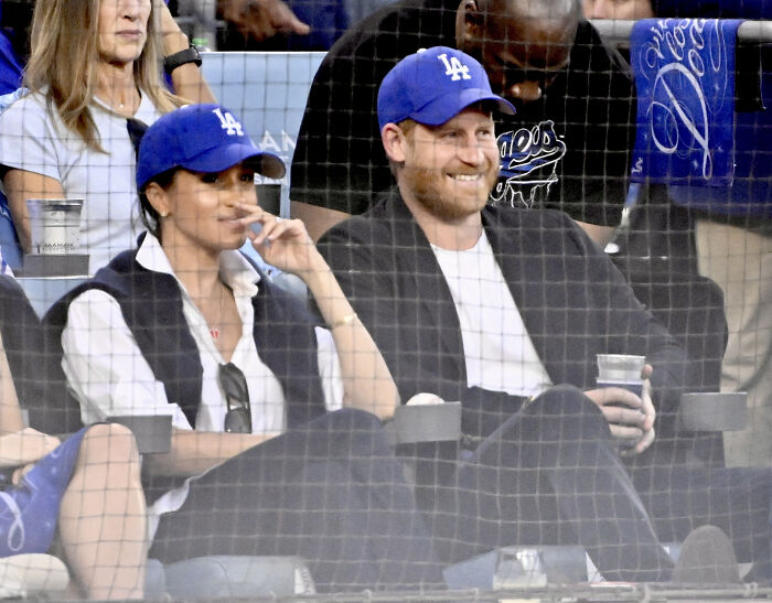 Meghan Markle and Prince Harry wearing blue hats, sitting together at a baseball game, enjoying a staged celebration. Meghan Markle and Prince Harry wearing blue hats, sitting together at a baseball game, enjoying a staged celebration.