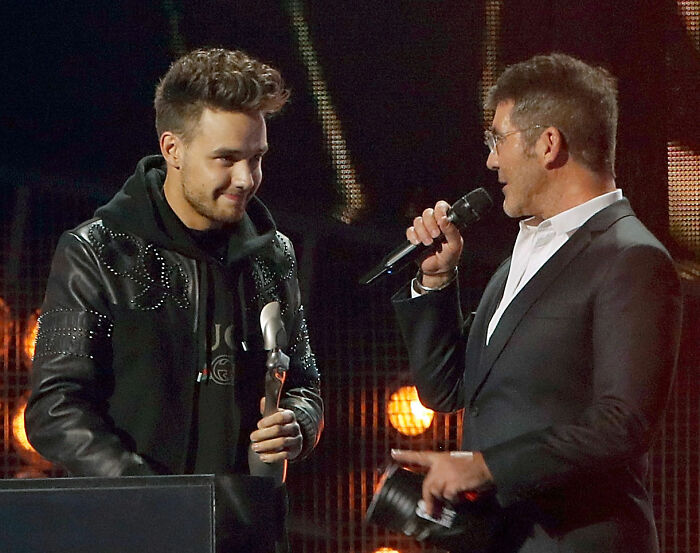 Simon Cowell speaking into a microphone while Liam Payne listens during a public event on stage. Simon Cowell speaking into a microphone while Liam Payne listens during a public event on stage.