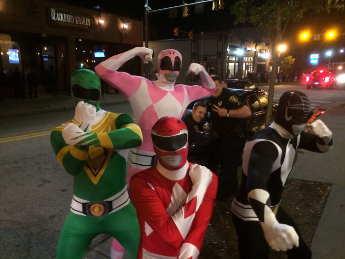 Four people in colorful superhero costumes pose on a street while police officers photobomb the scene in a humorous moment.