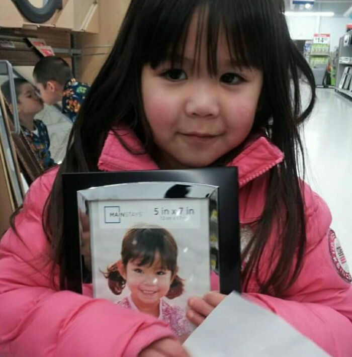 Young girl in a pink coat holding a photo frame with a photobomb in the background creating a funny moment.