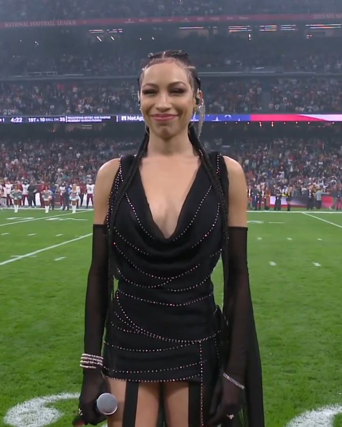 Singer wearing a black dress and gloves on the football field, criticized for disrespectful outfit during US National Anthem at NFL game.
