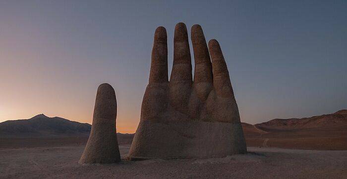Massive hand sculpture rising from desert sand at sunset, an iconic spectacular sculpture for global travel bucket lists.