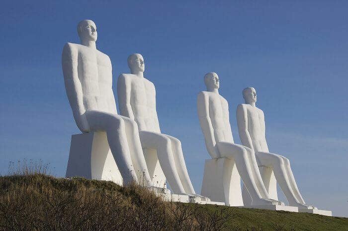 Four large white statues of seated men on a hill under clear blue sky, a spectacular sculpture featured worldwide.