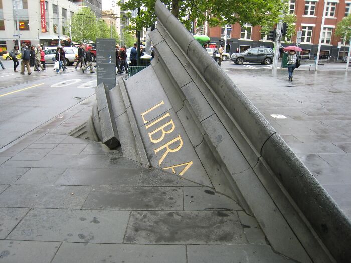 Modern outdoor sculpture titled Libra partially embedded in urban pavement with pedestrians and buildings in the background